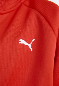 Tissu de sport texturé rouge avec un logo Puma blanc représentant un chat en train de sauter. Le design comprend un col côtelé et des accents aux épaules.
