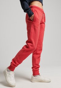 Person wearing red drawstring sweatpants and white sneakers, standing with one leg slightly bent and hands in pockets.