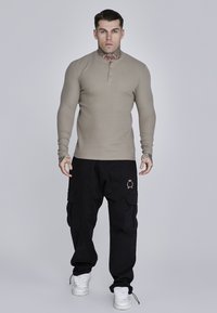 Bež majica dugih rukava henley s ovratnikom na gumbima, u kombinaciji s labavim crnim cargo hlačama s bočnim džepovima i grbom. Bijele tenisice.