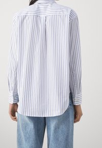 Bílá košile s tenkými modrými svislými pruhy, dlouhými rukávy s knoflíkovými manžetami, rovný zadní lem a texturou látky typickou pro bavlnu.