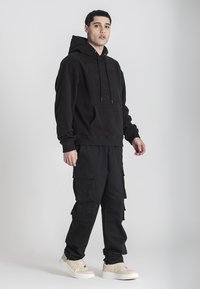 Czarna bawełniana bluza z kapturem z wypukłym logo niedźwiedzia, w zestawieniu z czarnymi spodniami cargo z wieloma kieszeniami i beżowymi sneakersami.