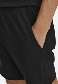 Pantalones cortos deportivos negros hechos de tejido texturizado con bolsillos frontales y una cinturilla elástica. Se pueden ver costuras detalladas a lo largo de las costuras.