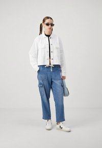 Veste en jean blanche raccourcie avec des boutons en bois, haut noir, pantalon cargo bleu et baskets blanches. Accessoires comprenant un sac à main gris et des lunettes de soleil.