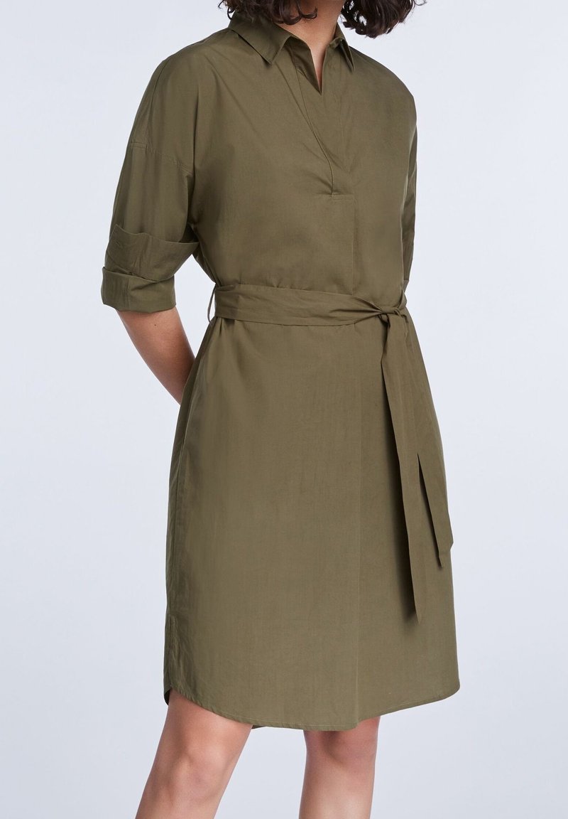 Femme portant une robe vert olive mi-longue avec manches mi-longues et une ceinture nouée à la taille, sur un fond clair uni.