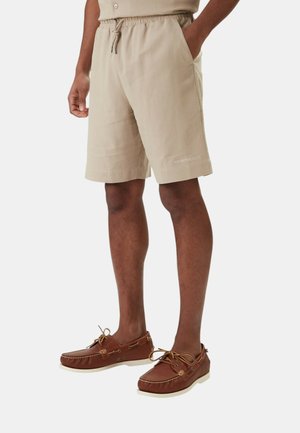 Uomo che indossa pantaloncini beige con coulisse e mocassini di pelle marroni, in piedi con una mano in tasca.