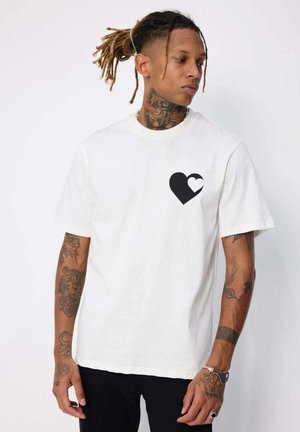 T-shirt en coton blanc avec un motif de cœur noir sur la poitrine gauche. Manches courtes, col rond et coupe décontractée.