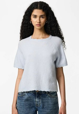 Jeune femme aux longs cheveux bouclés portant des boucles d'oreilles argentées en forme de créoles, un pull bleu clair à manches courtes et duveteux, et un jean en denim bleu, posant devant un fond blanc.