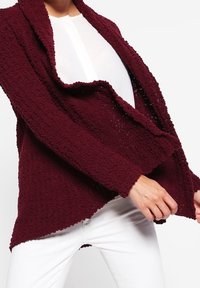 Bordeaux geklede cardigan met een open voorkant, gedrapeerd ontwerp en een oneven zoom; laag over een witte top, gedragen met witte broeken.