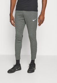 Pantalones deportivos grises con un diseño ajustado, que presentan un logo de swoosh blanco de Nike y una textura sutil. Combinados con zapatos negros.