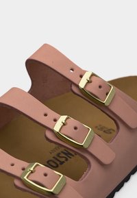 Ροζ λουράκια σανδαλιού με τρεις ρυθμιζόμενες αγκράφες από χρυσό μέταλλο πάνω σε ταμπά πάτο, που δείχνει μέρος της μαύρης σόλας από κάτω.