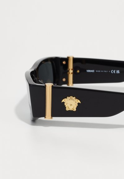 Black sunglasses with a thick frame, featuring gold accents and a decorative emblem on the side. Textured temples and tinted lenses.
