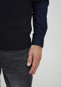 Czarny, dziergany sweter z prążkowanym dołem, noszony na ciemnoniebieskiej koszuli z długim rękawem. Widoczne szare dżinsy, pokazujące zbliżenie na dłoń spoczywającą obok.