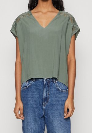 Person iført en løs grøn V-hals top med bløde skulderdetaljer i blonder og blå højtaljede denimjeans mod en ensfarvet baggrund.