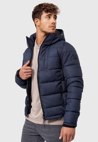Marineblå polstret jakke med lynlås foran og en hætte. Har elastiske manchetter, fleeceforing og et struktureret stof. Letvægtsdesign.