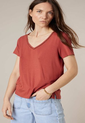 Femme portant un t-shirt à manches courtes couleur rouille avec un col en V et un jean bleu clair, mains dans la poche et bracelet en or au poignet.