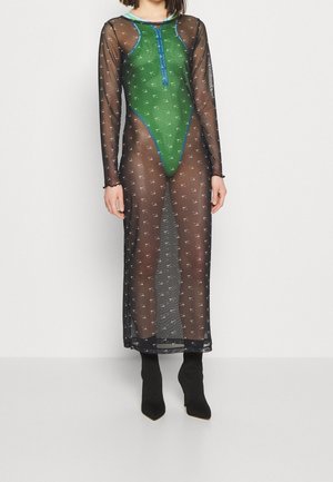 Femme portant un maillot de bain vert sous une longue robe noire en voile avec un petit motif répété, associée à des bottines noires à talons hauts.