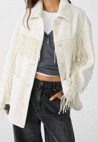Giacca bianca con frange realizzata in morbido tessuto, caratterizzata da bottoni beige, due tasche frontali e una vestibilità rilassata, abbinata a jeans neri a vita alta.