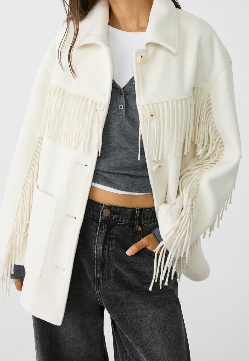 Giacca bianca con frange realizzata in morbido tessuto, caratterizzata da bottoni beige, due tasche frontali e una vestibilità rilassata, abbinata a jeans neri a vita alta.