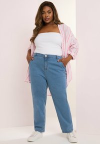 Camisa abotonada de rayas en rosa claro y blanco, superpuesta sobre una blusa blanca sin mangas, combinada con jeans de denim azules de talle alto y zapatillas blancas.