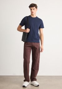 T-shirt a collochure navale, pantaloni larghi marroni con tasche, borsa a spalla nera e sneakers bianche su uno sfondo neutro.