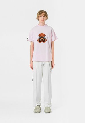 Camiseta de manga corta rosa con un gráfico de un oso marrón y negro, combinada con pantalones cargo blancos y zapatos verde claro texturizados.