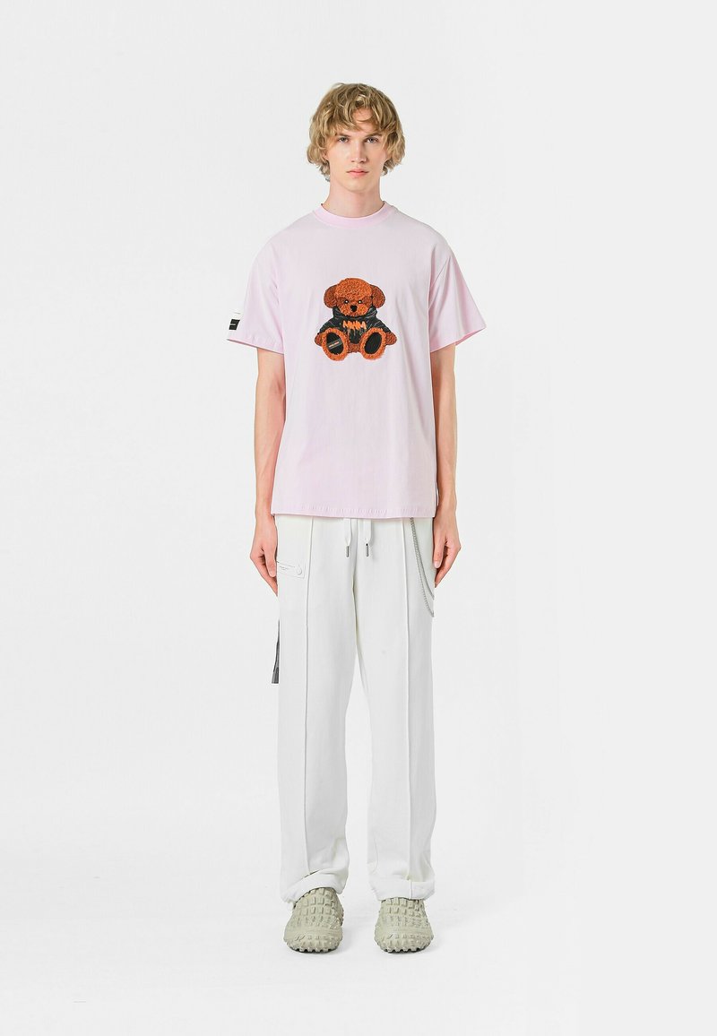 Camiseta de manga corta rosa con un gráfico de un oso marrón y negro, combinada con pantalones cargo blancos y zapatos verde claro texturizados.