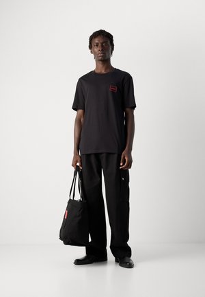 Mann in schwarzem Hugo-T-Shirt und weiten Hosen, der eine schwarze Tragetasche hält und vor einem schlichten weißen Hintergrund steht.