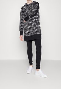 Czarna sukienka z kapturem, z powtarzającym się wzorem logo, długimi rękawami i kieszenią kangurką. Połączona z czarnymi legginsami i białymi sneakersami.