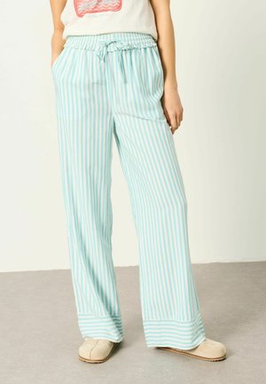 Frau trägt hellblaue und weiße vertikal gestreifte weit geschnittene Hose mit elastischem Bund und beigefarbenen Slip-On-Schuhen, steht auf grauem Boden.