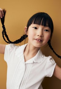 Jeune fille aux cheveux tressés portant un chemisier blanc, tenant ses tresses levées sur un fond jaune moutarde.