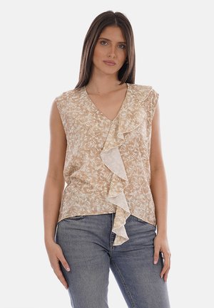 Donna con lunghi capelli castani che indossa un top beige senza maniche con stampa floreale e un volant frontale, abbinato a jeans blu, posando davanti a uno sfondo bianco.