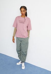 Svetlo roza kratkotrajna majica s kratkimi rokavi z napisom "New Balance 1906" v temno rjavi barvi, v kombinaciji z zelenimi cargo hlačami in belimi športnimi čevlji, pod njo pa je dolgosleeva majica s črtami.