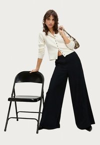 Cardigan bianco con bottoni, pantaloni neri a gamba larga a vita alta e borsa stampata, posati accanto a una sedia pieghevole nera su uno sfondo neutro.