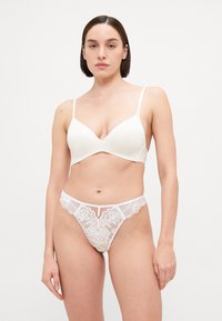 Weißes BH- und String-Set, bestehend aus glattem Stoff und zarter Spitzenverzierung. Der BH hat dünne Träger und ein V-Ausschnitt-Design; der String verfügt über florale Spitze.