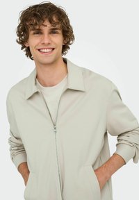 Veste zippée beige clair avec un col ouvert, poches latérales et manches longues. Texture lisse et design minimaliste.