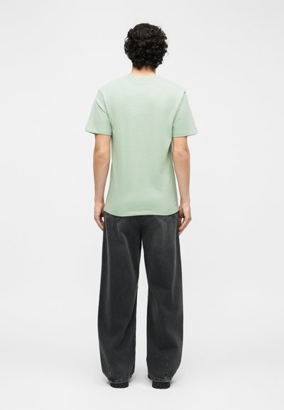 Άνδρας με σγουρά μαλλιά, που στέκεται με την πλάτη προς τα εμπρός, φοράει ανοιχτό πράσινο κοντομάνικο μπλουζάκι και φαρδιά μαύρα τζιν, μπροστά σε λευκό φόντο.