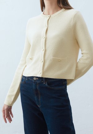 Femme portant un cardigan crème texturé boutonné avec deux petites poches avant, associé à un jean bleu foncé taille haute.