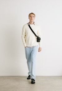 Suéter de canalé color crema, jeans azules claros y mocasines negros. La modelo lleva un pequeño bolso cruzado negro. Entorno minimalista con un fondo neutro.