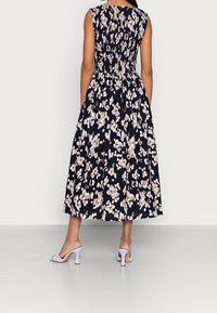Eine Person, die ein ärmelloses, blumengemustertes Kleid trägt, steht vor einem weißen Hintergrund und präsentiert gestrickte lila High Heels.