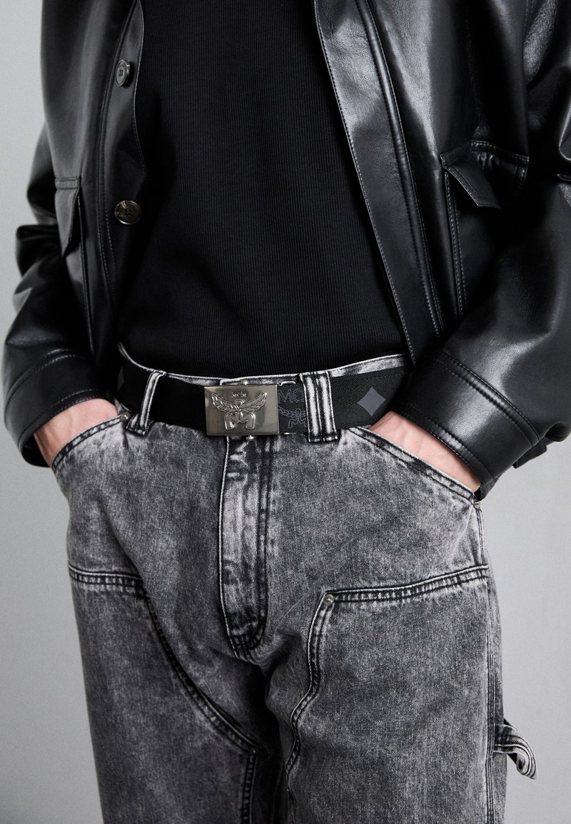 Chaqueta de cuero negra sobre una camiseta negra de canalé, combinada con pantalones de denim grises. Presenta un cinturón con hebilla plateada y detalles de costura prominentes.