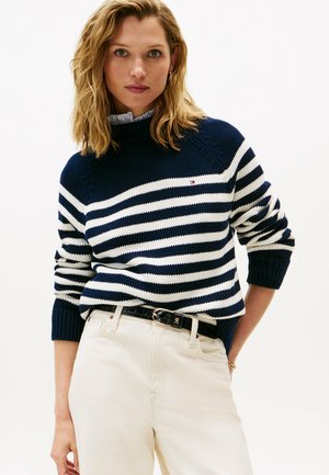 Woman with shoulder-length blonde hair wearing a navy and white striped sweater, tucked into high-waisted white pants with a black belt.
