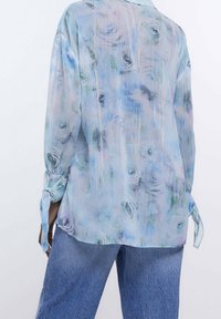 Light blue blouse with a floral print, featuring a textured fabric and tie details at the cuffs. Paired with denim jeans.