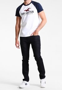 Camiseta blanca con mangas raglán azul marino y logotipo "HOLLISTER CALIFORNIA", combinada con vaqueros oscuros y zapatillas blancas. Corte y estilo casual.