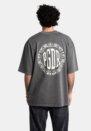 Overdimensineret grå t-shirt med korte ærmer, der har et hvidt cirkulært blomstermotiv og "PGDR" tekst på ryggen. 100 % bomuldsstof.