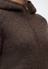 Gros plan d’une personne portant un sweat à capuche zippé marron tricoté avec une texture en laine et un tirette de fermeture éclair argentée.