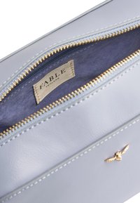 Borsa in pelle di colore grigio chiaro con zip e accessori dorati. L'interno presenta una fodera blu scuro e un'etichetta in tessuto con la scritta "FABLE ENGLAND".