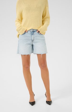Maglione lavorato a maglia giallo chiaro, pantaloni shorts in denim blu con orlo sfrangiato e tacchi a punta neri. I pantaloni shorts hanno una vestibilità casual.