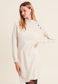 Robe en tricot crème avec des poignets côtelés, un col haut et des boutons d'épaule. Elle présente une coupe décontractée et une surface douce et texturée.