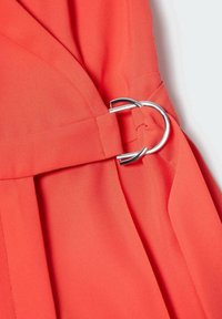 Robe corail avec une taille cintrée et une boucle métallique argentée. Le tissu est lisse, avec une texture légère et fluide, et des détails plissés.