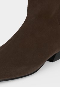 Gros plan d'une botte cheville en daim marron avec un petit talon bloc noir sur fond blanc.